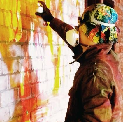 Person wearing a colorful cap and respirator spray-painting a brick wall with orange and pink graffiti on it.