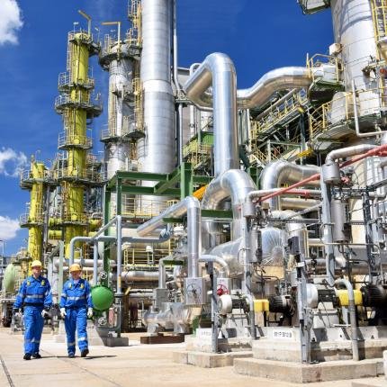 Two workers in blue coveralls and yellow hard hats walk beside a maze of silver industrial pipes at a chemical plant.